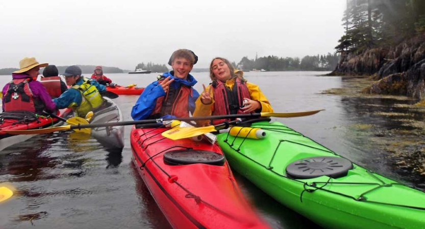 Two people wearing life jackets sit in two kayaks and give peace signs to the camera. 